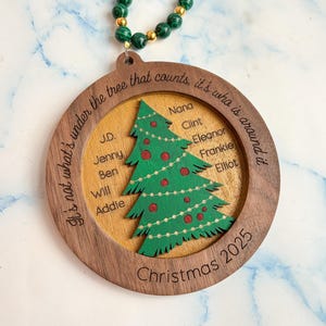 May include: Wooden Christmas ornament with a green Christmas tree design, red ornaments, and the names of family members. The ornament has a wooden frame with the text "It's not what's under the tree that counts, it's who is around it" and "Christmas 2025".