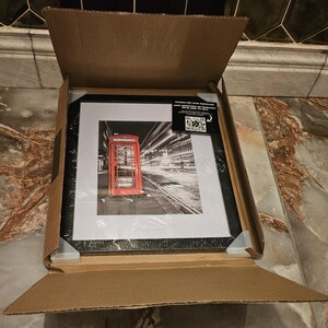 May include: A framed black and white photograph of a London telephone booth in a cardboard box. The red booth is the focal point, set against a blurred background of city lights. The frame is black, and the print is protected by clear plastic.