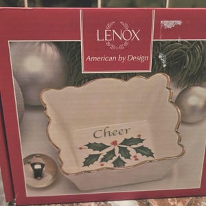 Puede incluir: Una caja roja que presenta un plato de cerámica blanco con borde festoneado y ribete dorado. El plato tiene la palabra "Cheer" y un diseño de acebo. La caja también muestra el logotipo de Lenox y la frase "American by Design."