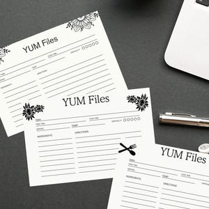 Può includere: Biglietti di ricette biancastri con il testo "YUM Files" e motivi floreali. Ogni biglietto ha sezioni per piatto, tempo di preparazione, ingredienti e istruzioni. Una tazza di caffè e un laptop sono sullo sfondo.