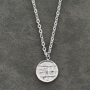 Könnte beinhalten: Silberfarbene Halskette mit rundem Anhänger. Der Anhänger hat ein strukturiertes, mosaikartiges Design. Die Kette ist eine filigrane Gliederkette, und der Anhänger hängt an einer kleinen Schlaufe. Der Hintergrund ist eine strukturierte graue Oberfläche.