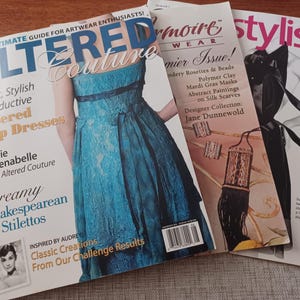 May include: Three fashion magazines are stacked on a wooden surface. The top magazine, "Altered Couture," features a teal dress. The middle magazine, "Drmoire Wear," has a black and gold illustration. The third magazine is titled "stylish."