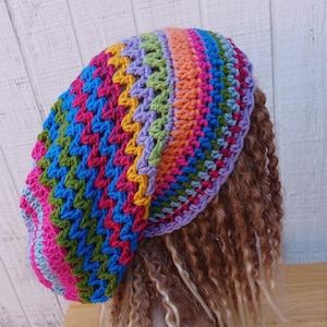 May include: A slouchy crochet beanie featuring a vibrant, multi-colored chevron pattern in shades of pink, blue, green, yellow, orange, and purple. The hat is crafted with a textured stitch design and displays a wide, rounded shape that sits loosely.