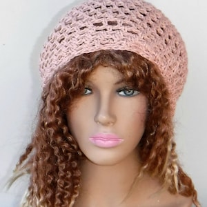 May include: A light pink crocheted hat with an open weave design. The hat is round and sits atop a mannequin head with long, curly brown and blonde hair. The hat appears to be handmade.