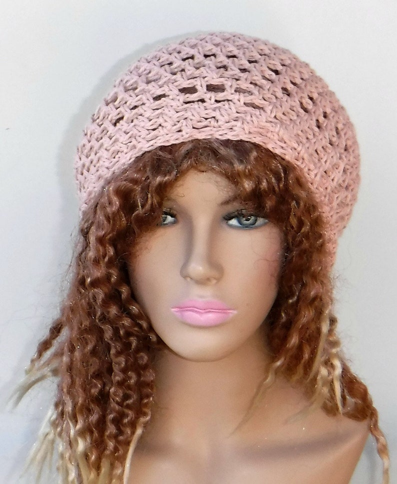 May include: A light pink crocheted hat with an open weave design. The hat is round and sits atop a mannequin head with long, curly brown and blonde hair. The hat appears to be handmade.