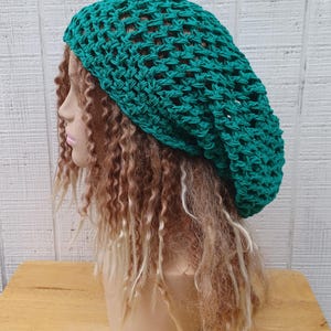 May include: A teal green crochet beanie hat with a loose, open weave. The hat is shown on a mannequin with long, curly hair.