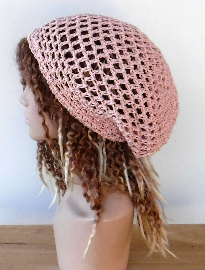 May include: A light pink crocheted hat with a net-like design. The hat is worn on a mannequin head with brown and blonde curly hair. The hat is a soft, neutral color.