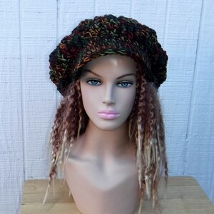 May include: A crocheted beanie in a multi-colored pattern of green, brown, orange, and red. The beanie has a slouchy, relaxed fit and is worn on a mannequin with long, curly hair.