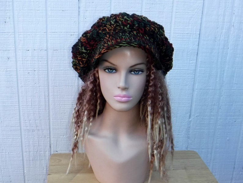 May include: A crocheted beanie in a multi-colored pattern of green, brown, orange, and red. The beanie has a slouchy, relaxed fit and is worn on a mannequin with long, curly hair.