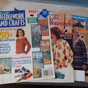 May include: A stack of vintage craft magazines, including "Needlework and Crafts" and "Call's Needlework." The magazines feature images of cross-stitch, knitting, and crochet projects, with text such as "50+ New Patterns" and "Autumn Sweaters to Knit & Crochet."