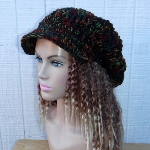 May include: A brown, green, and orange crocheted beanie with a wide brim. The beanie is worn on a mannequin with long, curly brown and blonde hair.