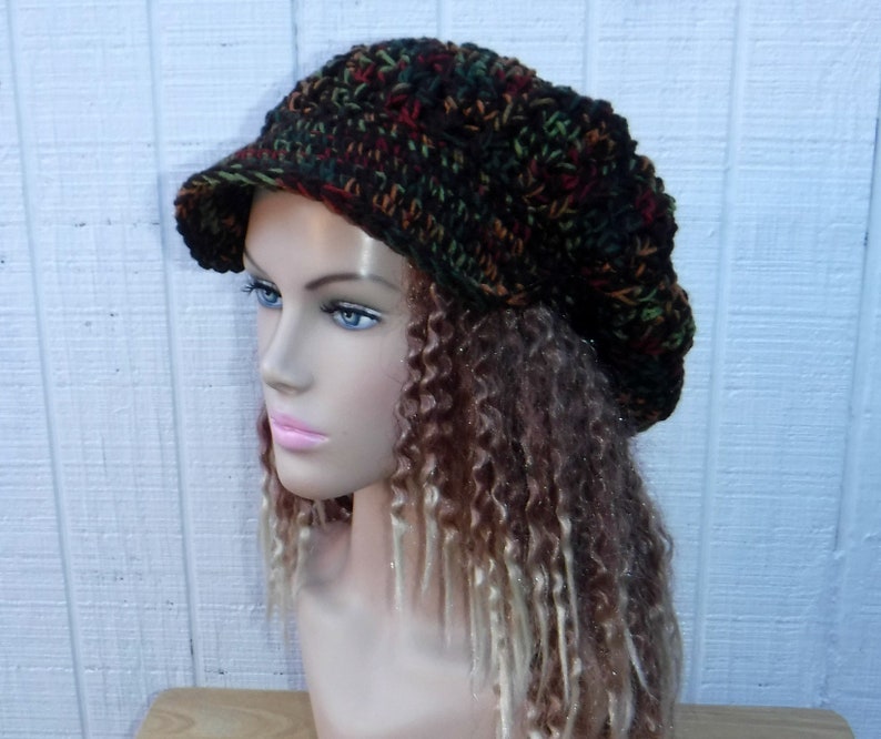 May include: A brown, green, and orange crocheted beanie with a wide brim. The beanie is worn on a mannequin with long, curly brown and blonde hair.