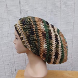 May include: A handmade, slouchy beanie in a camouflage pattern. The hat features a mix of green, brown, tan, and cream-colored yarn, creating a textured, striped design. The beanie is displayed on a mannequin head against a white wood-paneled background.