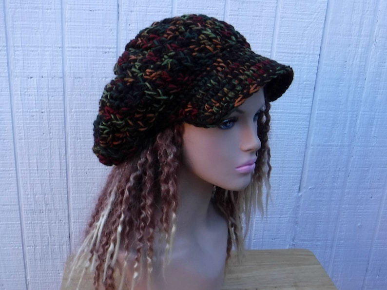 May include: A brown, green, and orange crocheted beanie with a wide brim. The beanie is worn on a mannequin with long, curly brown and blonde hair.