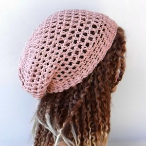 May include: A light pink, open-weave crochet beret. The hat is made of a soft, textured yarn and has a rounded crown and a close-fitting brim. The beret is shown on a person with long, curly brown hair.