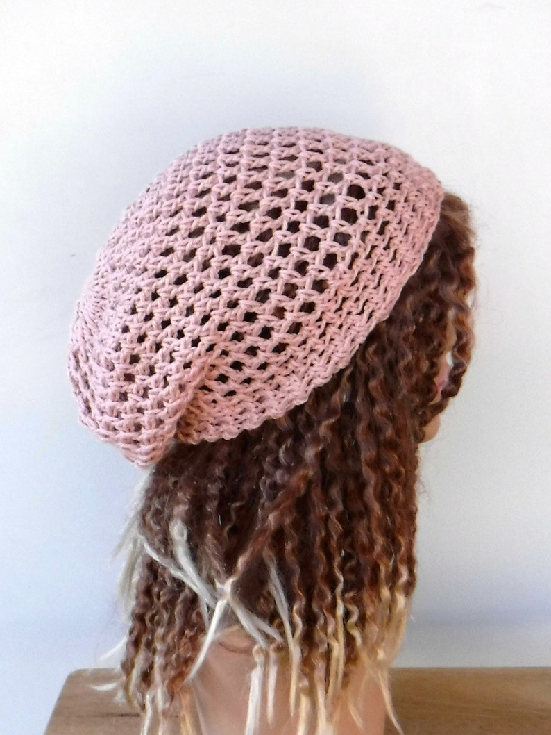 May include: A light pink, open-weave crochet beret. The hat is made of a soft, textured yarn and has a rounded crown and a close-fitting brim. The beret is shown on a person with long, curly brown hair.