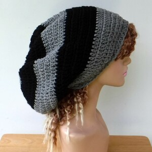 May include: A handmade crochet beanie in a striped pattern of black and gray. The hat is shown on a mannequin head with long, curly hair. The beanie is a slouchy style, suitable for casual wear.