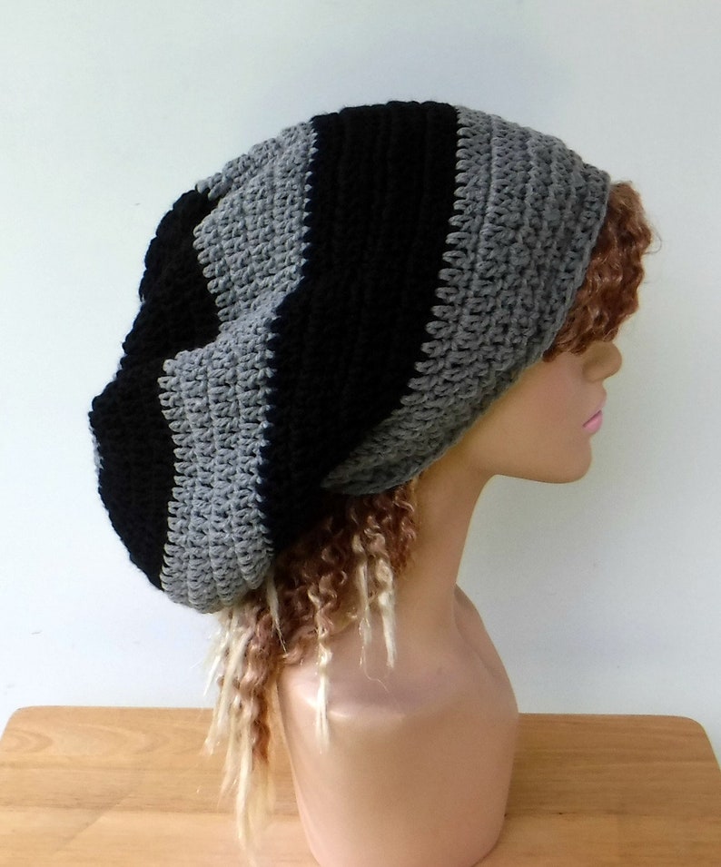 May include: A handmade crochet beanie in a striped pattern of black and gray. The hat is shown on a mannequin head with long, curly hair. The beanie is a slouchy style, suitable for casual wear.