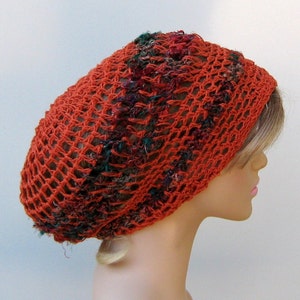 May include: A crocheted beanie hat in a burnt orange color with green and brown accents. The hat is made with a loose, open weave and has a slouchy fit.