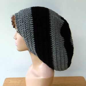 May include: A handmade crochet beanie in a striped pattern of black and gray. The hat is made of soft yarn and has a ribbed texture. The beanie is designed to be worn on the head.