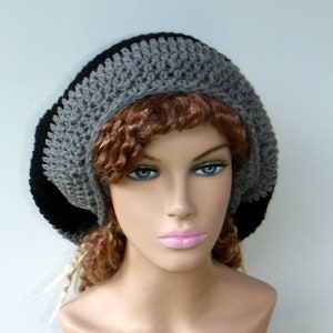 May include: A crocheted hat in a beret style. The hat features a color block design with a black brim, a gray middle section, and a black top. The hat is made of a soft, textured yarn.
