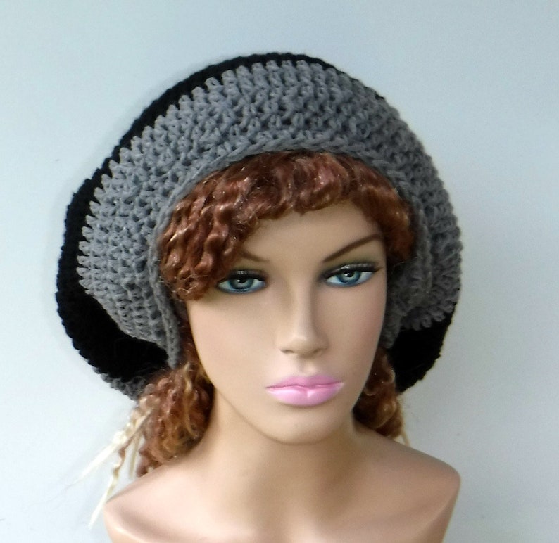 May include: A crocheted hat in a beret style. The hat features a color block design with a black brim, a gray middle section, and a black top. The hat is made of a soft, textured yarn.