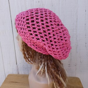 May include: A pink crocheted beanie hat with a loose, open weave. The hat is worn on a mannequin with long, brown dreadlocks.