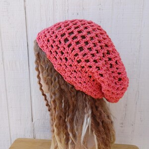 May include: A coral pink crocheted beanie hat with a loose, slouchy fit. The hat is made with a simple, open stitch pattern.