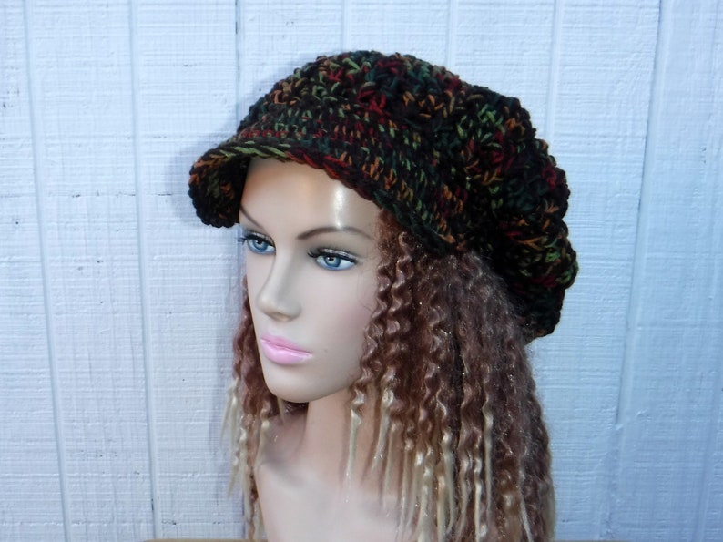 May include: A brown, green, and red crocheted beanie with a brim. The beanie is worn on a mannequin with long, brown and blonde curly hair.