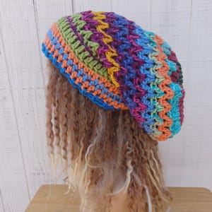 May include: A slouchy crochet beanie featuring a vibrant, multi-colored striped pattern in shades of blue, orange, green, purple, and yellow. The hat is constructed with a textured stitch pattern and sits atop a mannequin head with long, wavy hair.