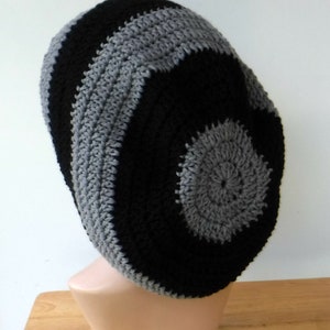 May include: A black and gray crocheted beanie hat. The hat features a spiral design with alternating bands of black and gray yarn. The hat is displayed on a mannequin head, showcasing its shape and texture. The hat is a handmade item.