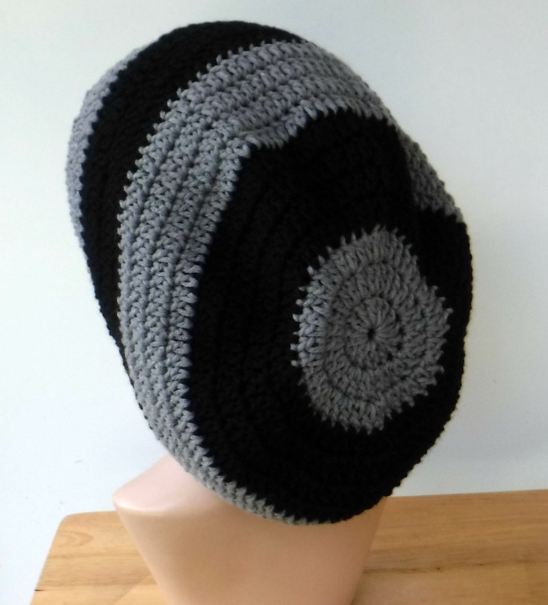 May include: A black and gray crocheted beanie hat. The hat features a spiral design with alternating bands of black and gray yarn. The hat is displayed on a mannequin head, showcasing its shape and texture. The hat is a handmade item.