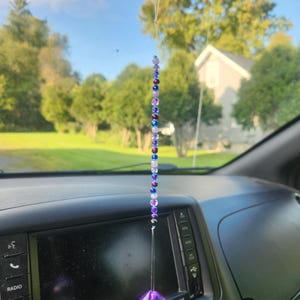 May include: A purple crystal suncatcher hanging from the rear view mirror of a car. The suncatcher is made of beads in shades of purple, blue and white.