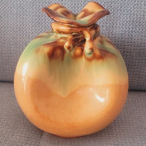 May include: A ceramic vase with a rounded body and a unique top design. The vase features a gradient of colours, transitioning from a light green at the top to a warm orange at the base. The top has a knotted design.