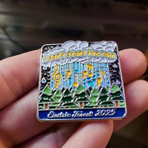 May include: Square enamel pin with a blue border featuring the text "I can't stand the rain" above a musical staff with notes and rain. Below are green trees and the text "Electric Forest 2025".