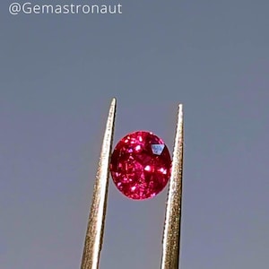 Peut inclure: Une pierre précieuse ovale rouge vif tenue par des pinces sur un fond gris.