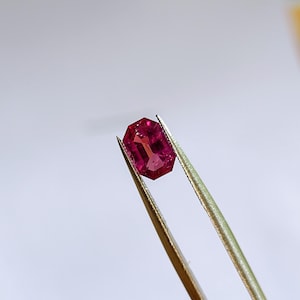 May include: A vibrant, rectangular-cut, deep red gemstone is held by tweezers against a white backdrop. The gem has a polished surface and reflects light, showcasing its internal structure. This gemstone is likely a ruby, suitable for jewellery.