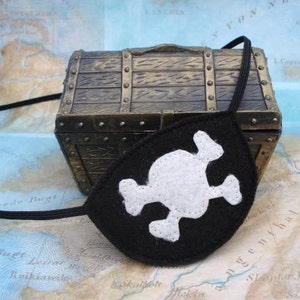 May include: A black felt pirate eye patch with a white skull and crossbones design. The eye patch is lying on a map with a small wooden treasure chest.