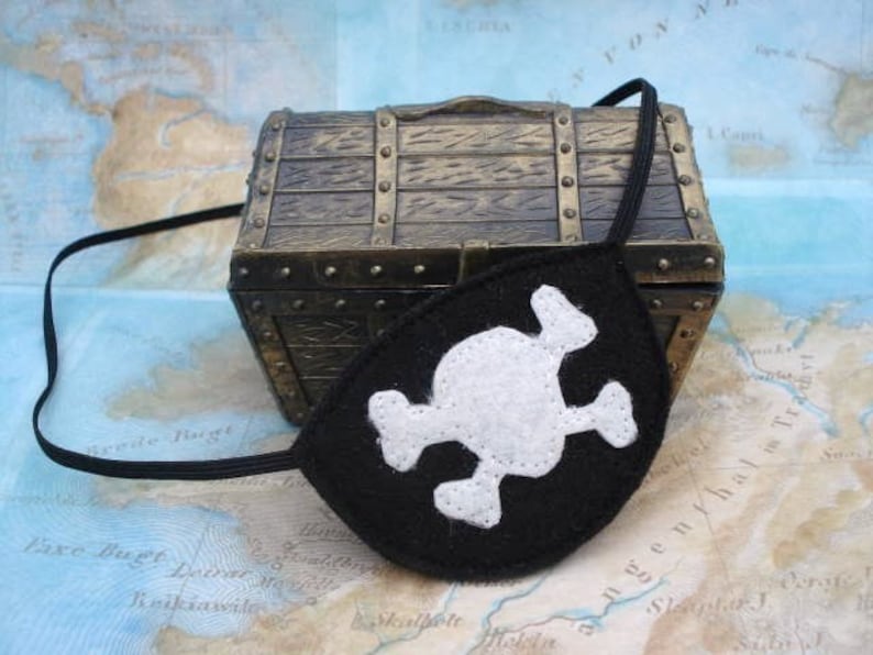 May include: A black felt pirate eye patch with a white skull and crossbones design. The eye patch is lying on a map with a small wooden treasure chest.