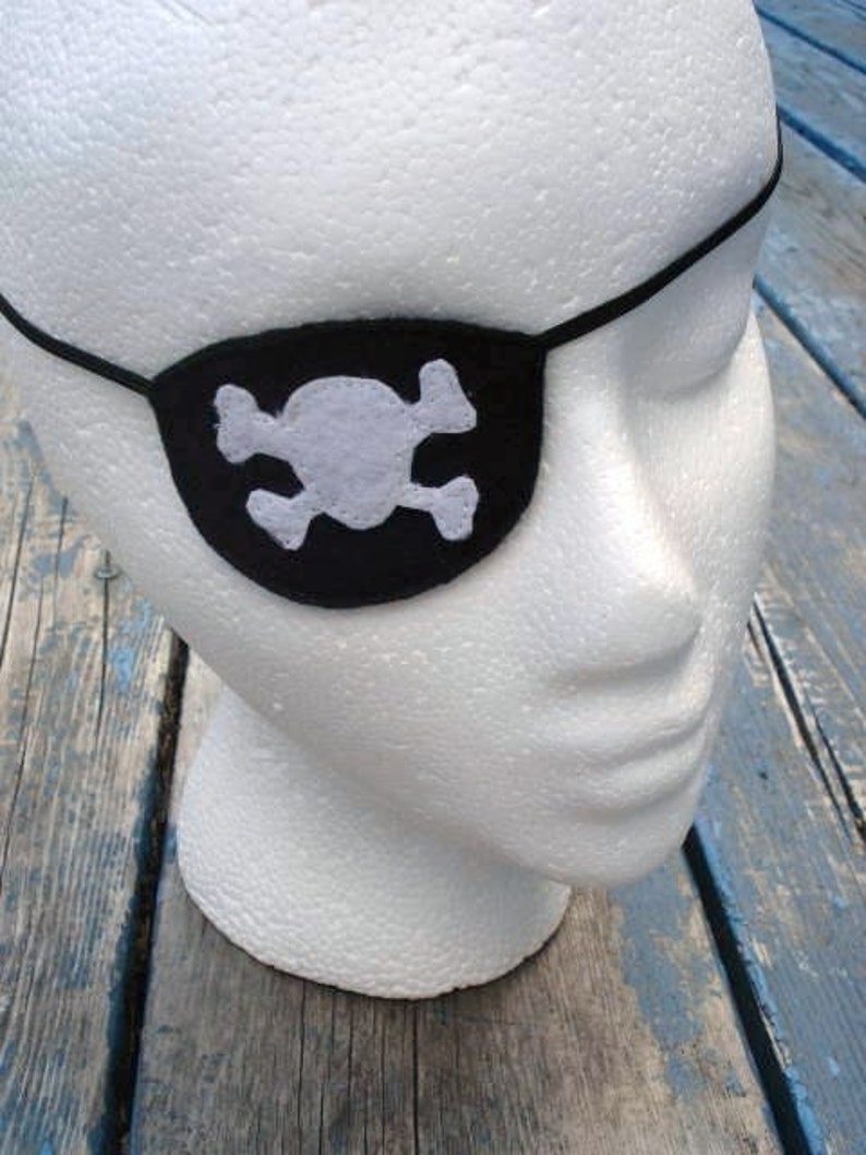 May include: A white foam mannequin head wearing a black felt pirate eye patch with a white felt skull and crossbones design.