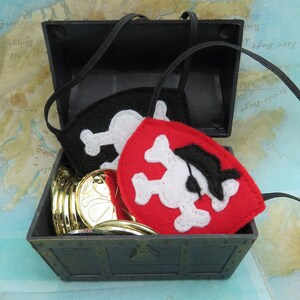 May include: A black plastic treasure chest with a gold coin design on the front. The chest is open and contains a stack of gold coins and two felt pirate eye patches. One eye patch is black with a white skull and crossbones, and the other is red with a white skull and crossbones and a black eye patch.