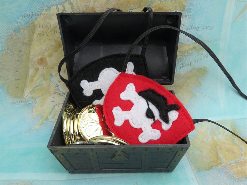 May include: A black plastic treasure chest with a gold coin design on the front. The chest is open and contains a stack of gold coins and two felt pirate eye patches. One eye patch is black with a white skull and crossbones, and the other is red with a white skull and crossbones and a black eye patch.