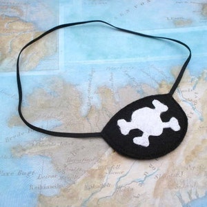 May include: A black felt pirate eye patch with a white skull and crossbones design. The eye patch is attached to a black elastic band.
