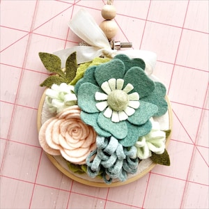 May include: A felt flower hoop with a green, white, and peach flower arrangement. The hoop is made of wood and has a light brown finish.
