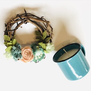 May include: A wreath made of twigs with felt flowers and greenery. The wreath is next to a teal blue enamel mug with a white candle inside.
