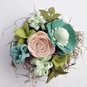 May include: A felt flower arrangement in a nest of twigs. The arrangement features a peach-colored rose, a teal flower with a light green center, and several green and light green felt leaves.