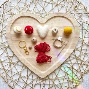 May include: A heart-shaped wooden tray holds craft supplies. There are various colorful felt balls, wooden beads, a gold keychain, red yarn, a needle, and a spool of thread. The tray sits on a gold geometric patterned surface.