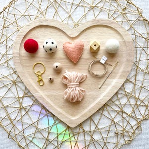 May include: A heart-shaped wooden tray holds craft supplies. A red felt ball, a black and white spotted ball, a pink felt heart, wooden beads, a gold keychain, twine, a needle, and a bundle of pink yarn are arranged on the tray.
