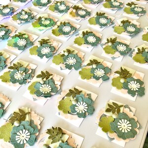 May include: A collection of felt flower embellishments in various shades of green, white, and cream. The flowers are arranged on a white surface and are ready to be used for crafting projects.