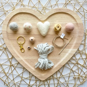 May include: A heart-shaped wooden tray holds craft supplies. Items include felt balls, a heart, a wooden geometric shape, a gold keychain, wooden beads, gray yarn, twine, and a needle. The tray sits on a gold geometric patterned surface.